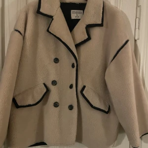 Oversize beige teddyjacka Stories S - Säljer en beige teddyjacka från Stories i storlek S. Jackan har svarta kontrastdetaljer längs kanterna, dubbelknäppning med svarta knappar och två fickor framtill. Oversized passform och mjukt/fluffigt material. 