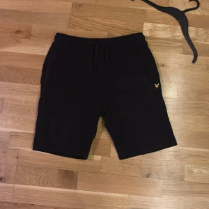 Svarta shorts från Lyle & Scott - Svarta shorts från Lyle & Scott med elastisk midja och snörning. Klassisk design med fickor på sidorna och en bakficka. Liten gul logga broderad framtill. Perfekta för en avslappnad stil.