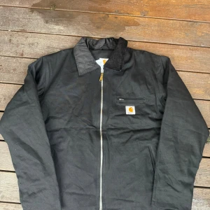 Svart Carhartt overshirt med dragkedja - Svart overshirt från Carhartt med klassisk krage och dragkedja framtill. Jackan har en bröstficka med Carhartt-logga och två sidofickor. Materialet är robust bomull med polyesterfoder, perfekt för lager-på-lager. Enkel och stilren design som funkar till streetwear.
