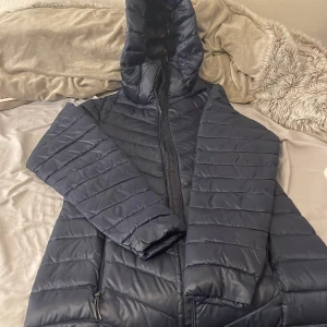 Mörkblå pufferjacka från McKinley - Snygg mörkblå pufferjacka från McKinley i storlek 36. Jackan har huva, dragkedja framtill och praktiska fickor med dragkedja. Quiltad design och lätt, syntetmaterial som håller dig varm. Perfekt för kyliga dagar och enkel att matcha med din stil.