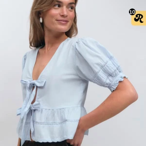 Ljusblå blus med knyt från Gina Tricot - Supersöt ljusblå blus med puffiga korta ärmar, v-ringning och två knyt framtill. Blusen har volangdetaljer och spetsinslag på ärmar och nederkant. Perfekt för dig som gillar romantisk och feminin stil. Materialet är mjuk bomull. Aldrig använd. Nypris 400kr.