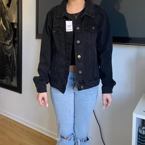 Svart jeansjacka från Boohoo - Svart jeansjacka från Boohoo med klassisk krage, raka axlar och knappar framtill. Jackan har två bröstfickor med lock och långa ärmar med knapp vid mudden. Snygg loose passform som funkar till många olika stilar. Aldrig använd, pris kan diskuteras 
