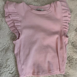 Rosa ribbad topp med volang från Zara - Supersöt ljusrosa topp från Zara i ribbad bomull med volangdetaljer på axlarna. Modellen är ärmlös och har rund halsringning. Perfekt för dig som gillar en feminin och trendig look. Säljes i storlek S.