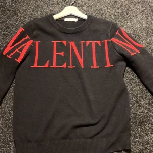 Svart stickad tröja från Valentino - Svart stickad tröja från Valentino med stor röd logotyp över bröstet och ärmarna. Tröjan har rund halsringning och ribbade muddar vid ärmslut och nederkant. Snygg och statement med kontrasterande färger.