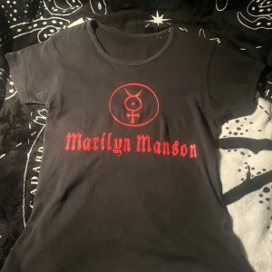 Svart Marilyn Manson t-shirt S/M - Svart t-shirt med rött Marilyn Manson-tryck och symbol på bröstet. Klassisk rund hals och korta ärmar. Perfekt för dig som gillar bandmerch och vill sticka ut med en edgy stil.