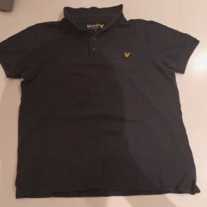 Mörkblå pikétröja från Lyle & Scott - Snygg mörkblå pikétröja från Lyle & Scott med klassisk krage, korta ärmar och knäppning framtill. Tröjan har det ikoniska gula logobroderiet på bröstet och är tillverkad i mjuk bomull. Perfekt för en clean och stilren look.