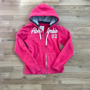 Rosa hoodie från Abercrombie & Fitch - Snygg rosa hoodie från Abercrombie & Fitch med broderad logga och siffran 92 på bröstet. Tröjan har huva med dragsko, dragkedja framtill och mjukt fodrad insida. Perfekt för chill dagar och streetstyle.  Bilder på måtten finns tillgängliga. Fråga gärna! Snabb leverans!
