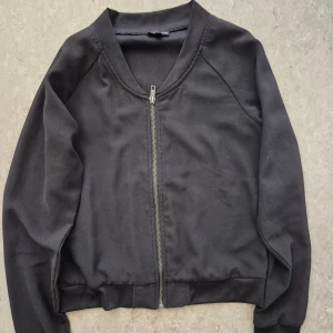 Jacka/Kofta - Svart jacka/kofta från KappAhl i storlek 158/164. Jackan har en klassisk rak passform, rundad krage och dragkedja framtill. Tillverkad i mjukt syntetmaterial med ribbade muddar vid ärmslut och nederkant. Perfekt för en chill och stilren look. Har en liten fläck.