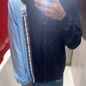 Lacoste tvåfärgad vindjacka - Säljer en riktigt snygg vindjacka från Lacoste med cool tvåfärgad design i ljusblått och mörkblått. Jackan har huva, dragkedja och elastiska muddar. På framsidan finns ett vitt band med Lacoste-loggor och den klassiska krokodilen på bröstet. Köpt på Lacoste store för 1800 kr den är i ny skick då den bara är använd en gång ute 2 gånger inomhus säljs för att dene lite för stor för mig pris kan diskuteras 