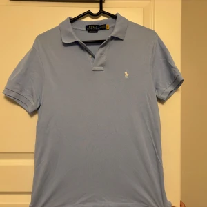 Ljusblå Polo Ralph Lauren pike  - Snygg ljusblå t-shirt från Polo Ralph Lauren med klassisk krage och två knappar vid halsen. Custom slim fit-modell med korta ärmar och diskret broderad logga på bröstet. Perfekt för dig som gillar stilrena och tidlösa plagg.