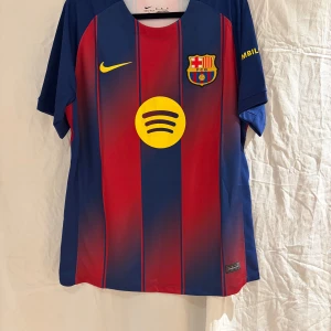 FC Barcelona Lamine Yamal tröja Nike M -  En  FC Barcelona fotbollströja  i storlek M med Lamine Yamal och nummer 10 på ryggen. Tröjan har klassiska blå och röda vertikala ränder, gult tryck och klubbmärke på bröstet. Tillverkad i lätt Dri-FIT material som andas.