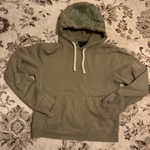 Syna world  - Olivgrön hoodie från SNK med fet graffiti-print på huvan och grova vita snören. Tröjan har känguruficka framtill och mjukt material som känns chill. Perfekt för dig som gillar streetwear och vill sticka ut lite extra.