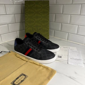 Gucci ace svarta sneakers med röd/blå detalj - Gucci sneakers i svart med monogrammönster och vita platta sulor. Sidorna har klassiska röd-blå ränder och hälen har metallicdetaljer i rött och blått med Gucci-logga. Skorna har snörning och rund tå, tillverkade i skinn och syntetmaterial.