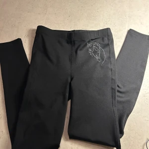 Svarta ridleggings  - Svarta ridleggings från CRW i stretchigt material, dekorerade med glittriga stjärnor längs benen och ett hästmotiv i silverprickar vid höften. Sitter tight och har elastisk midja för skön passform. Perfekta för dig som gillar ridning.