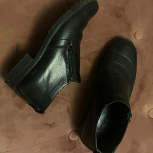 Svarta boots i skinn med dragkedja - Säljer ett par svarta boots i skinn med låg klack och rund tå. Skorna har dragkedja på sidan för enkel påtagning och en stilren, klassisk look som passar till många outfits. Perfekta för dig som gillar minimalistisk stil och vill ha ett par mångsidiga boots.