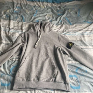 Grå Stone Island hoodie med patch - Säljer en grå hoodie från Stone Island med klassisk patch på vänster ärm. Tröjan har huva med snörning, känguruficka och ribbade muddar. Perfekt för dig som gillar streetwear och vill ha en clean look med ikonisk detalj. Storlek L men passar som M kom med bud och frågor 