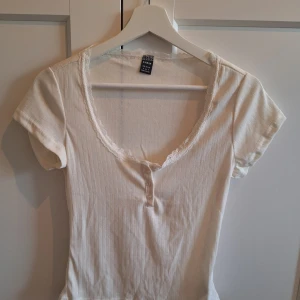 Vit ribbad topp med spets från SHEIN - Säljer en vit ribbad topp från SHEIN i storlek S. Toppen har korta ärmar, rundad halsringning med spetskant och tre små knappar framtill. Materialet är mjukt och stretchigt, perfekt för en chill och snygg look. Passformen är normal och toppen har diskreta vertikala ränder.
