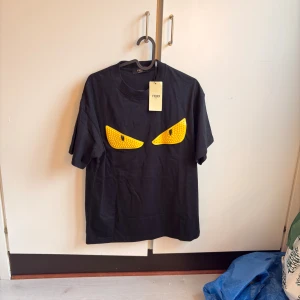 Fendi tshirt  - Cool svart t-shirt från Fendi med ikoniska gula ögon framtill. T-shirten har rund hals och korta ärmar, tillverkad i mjuk bomull. Perfekt för dig som vill sticka ut med en unik designerpiece och addera lite edge till din stil.                                           Storlek: M