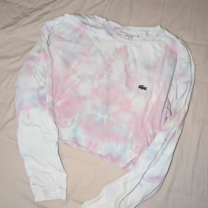 Pastell tie-dye croppad tröja Lacoste - Säljer en croppad långärmad tröja från Lacoste med pastellfärgat tie-dye mönster i rosa, blått och vitt. Tröjan har klassisk rund hals och den ikoniska Lacoste-loggan på bröstet. Perfekt för dig som gillar färg och chill vibe.