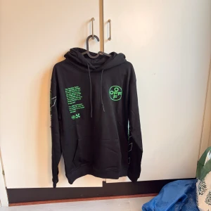  Off White hoodie  - Svart hoodie från Off-White med gröna grafiska tryck och text både fram och bak. Tröjan har huva med snörning, ribbade muddar och stor Off-White-logga på ryggen. Materialet är mjuk bomull och passformen är loose. Perfekt för dig som gillar streetwear och statement plagg.                          Storlek S 