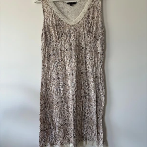 Beige ärmlös klänning från Twilfit - Snygg ärmlös klänning från Twilfit i beige och bruna toner med ett coolt ormmönster. Klänningen har v-ringning med spetskant och är gjord i mjuk modalblandning med stretch. Perfekt för dig som gillar unika mönster och bekväma material.