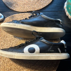 Svarta sneakers från Oliver Cabell - Snygga svarta sneakers från Oliver Cabell med vit sula och ett stort vitt cirkelmönster på sidan. Skorna har snörning och är tillverkade i skinn med rund tå och platt sula. Perfekta för dig som gillar stilrena och moderna detaljer.