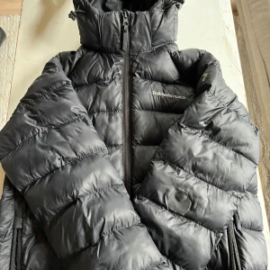 Svart Peak Performance pufferjacka  - Svart vinterjacka från Peak Performance med huva och dragkedja framtill. Jackan har quiltad design och är tillverkad i syntetmaterial. Snygg, varm och perfekt för kalla dagar. Klassisk logga på bröstet och praktiska fickor med dragkedja. Den har en lagning (se bild)