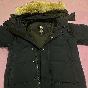 Mörkblå dunjacka Canada Goose - Säljer en mörkblå dunjacka från Canada Goose med fluffig beige päls på huvan. Jackan har dragkedja och knappar framtill, stora fickor och en klassisk, varm design som är perfekt för kalla dagar. Märket syns tydligt på insidan av jackan.