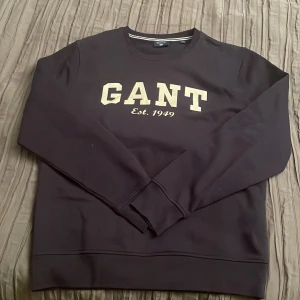 Marinblå sweatshirt från GANT i storlek L - Marinblå sweatshirt från GANT med stor beige logotyp och texten 'Est. 1949' på bröstet. Tröjan har rund hals, långa ärmar och ribbade muddar. Materialet känns mjukt och har en klassisk, stilren look som passar perfekt till jeans eller chinos.