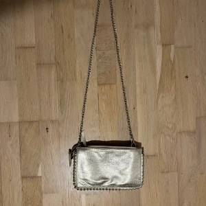 Guldig axelväska med kedja - Snygg guldig axelväska med kedjerem och dekorativa nitar längs kanten. Perfekt storlek för det viktigaste och ger en cool touch till din outfit. Väskan har dragkedja upptill och en stilren, glansig finish.