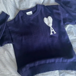Mörkblå stickad tröja från Ami Paris - Snygg mörkblå stickad tröja från Ami Paris med vit hjärta och A på bröstet. Tröjan har rund hals, ribbade muddar och är långärmad. Perfekt för dig som gillar stilrena detaljer och vill ha något som sticker ut.