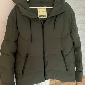 Grön pufferjacka från Woodbird - Säljer en grön pufferjacka från Woodbird med huva och svarta dragkedjor. Jackan har en clean look med raka linjer och två snedställda fickor framtill. Orginal pris 2399 nyskick nästintill oanvänd