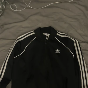 Svart Adidas kofta - Svart Adidas kofta med klassiska vita ränder längs ärmarna och vit piping. Jackan har dragkedja framtill, ribbad krage och Adidas-logga på bröstet. Perfekt för dig som gillar sportig och clean stil.