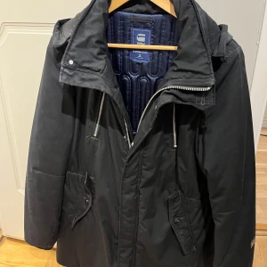 Svart parkajacka från G-Star RAW - Mycket fin svart parkajacka från G-Star RAW Women med huva, dragkedja och knappar framtill. Jackan har två stora fickor med lock och snygga detaljer på ärmen. Fodret är quiltat och jackan har en klassisk passform som är perfekt för kyliga dagar. Jackan är knappt använd och i nyskick!