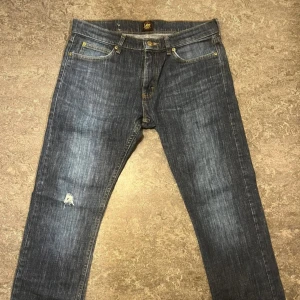  jeans från Lee, mörkblå  - Jeans från Lee i mörkblå tvätt med snygga slitningar och klassiska fem fickor. Jeansen har gul kontrastsöm, Lee-logga på knapp och bakficka samt en liten reva på vänster ben. Perfekt passform för dig som gillar smalare siluett.