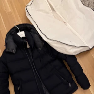 Svart Moncler pufferjacka med huva - Svart pufferjacka från Moncler med avtagbar huva, två sidofickor med dragkedja och logotyp på ärmen. Jackan har quiltad design och är fodrad med grått innerfoder. Perfekt för kalla dagar och har en snygg, modern look. Storlek 2