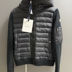 Svart pufferjacka från Moncler - Svart pufferjacka från Moncler med stickade ärmar och huva. Jackan har tvåvägsdragkedja, ribbad mudd och ficka med dragkedja på ärmen. Materialmix av polyester och stickad ull ger en schysst kontrast. Ikonisk Moncler-logga på ärmen. Kort vid midjan och lite tajt vid armarna