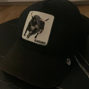 Svart keps Goorin Bros The Farm Raging Bull - Svart keps från Goorin Bros The Farm med broderad patch framtill som visar en tjur och texten 'RAGING'. Kepsen har mesh baktill för extra ventilation och justerbar snapback. Perfekt för dig som vill sticka ut med en cool accessoar. OBS det svarta som är i kepsen är ett tidigare namn men syns inte utifrån.