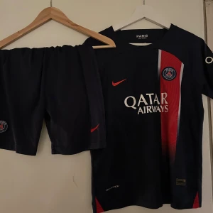PSG fotbollströja och shorts Nike - Säljer ett PSG matchställ från Nike med kortärmad tröja och shorts. Tröjan är marinblå med röd och vit rand längs framsidan, PSG-logga på bröstet och Qatar Airways tryck. Materialet är lätt och andas, perfekt för träning eller match.