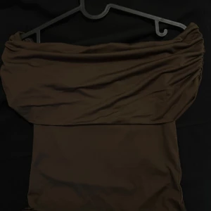 Brun offshoulder kortklänning - Snygg brun kortklänning med offshoulder-design och lätt rynkade detaljer vid axlarna. Klänningen har en figurnära passform och är tillverkad i ett mjukt, stretchigt material som sitter skönt mot kroppen. Perfekt för dig som gillar stilrena och trendiga plagg. Har använt den bara 1 gång 