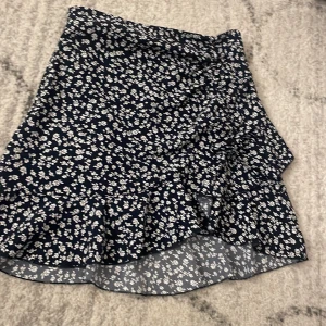 Blommig omlottkjol i marinblått - Supersöt kort omlottkjol i marinblått med vitt blommönster och volangkant. Perfekt för dig som gillar en feminin och trendig stil. Kjolen har en lätt och luftig känsla, vilket gör den skön att bära under varmare dagar.