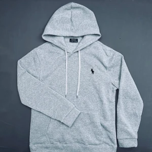 Grå hoodie från Polo Ralph Lauren - Snygg ljusgrå hoodie från Polo Ralph Lauren med klassisk broderad logga på bröstet. Tröjan har huva med snörning, känguruficka och långa ärmar med ribbade muddar. Perfekt för chill dagar och enkel att matcha med jeans eller joggers.