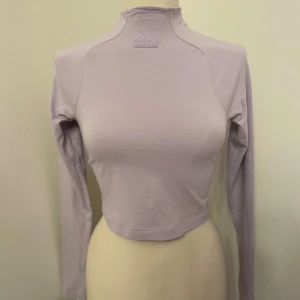 Ljuslila croppad långärmad topp Adidas - Säljer en ljuslila croppad långärmad topp från Adidas med hög hals och diskret logga framtill. Toppen har en slim passform och är tillverkad i mjukt material, perfekt för en sportig och trendig look. Har inte kommit till användning 