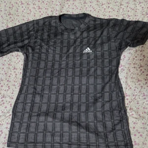 Svart rutig Adidas t-shirt - Snygg svart t-shirt från Adidas med vit logga på bröstet. T-shirten har ett rutigt mönster över hela plagget och klassisk rund hals. Materialet känns lätt och passar perfekt till sport eller chill. Kortärmad modell med normal passform.