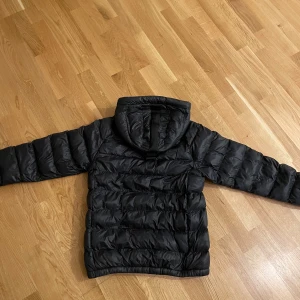 Svart dunjacka från Peak Performance - Säljer en svart dunjacka från Peak Performance med huva och dragkedja framtill. Jackan har en puffig look, ribbade muddar vid ärmslut och två sidofickor med dragkedja. Perfekt för kalla dagar. 