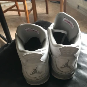 Nike Air Jordan 4 Retro vita sneakers - Nike Air Jordan 4 Retro sneakers i vitt med grå och svarta detaljer. Skorna har klassisk meshpanel på sidorna, synlig Air-sula och Jumpman-logga i rött på plösen och hälen. Tillverkade i skinn med snörning och platt sula. Perfekt för dig som gillar streetwear och ikoniska modeller.