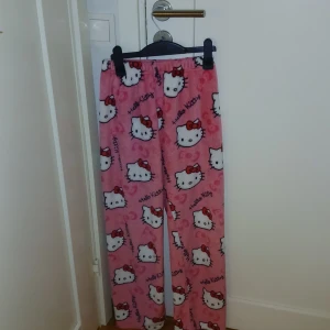 Hello Kitty rosa mjukisbyxor - Supermysiga rosa mjukisbyxor med Hello Kitty-tryck över hela byxorna. Byxorna har elastisk midja och är gjorda i mjukt fleece-material, perfekta för chill hemma. Söt design med röda rosetter och hjärtan som detaljer.