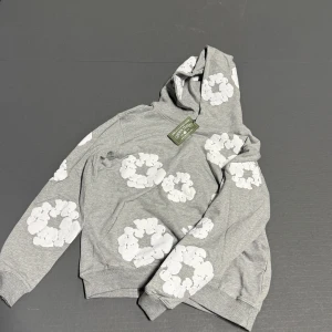 Grå hoodie med vita blommor Denim Tears - Grå hoodie från Denim Tears med stora vita blommor tryckta över hela tröjan. Tröjan har huva och långa ärmar, samt ribbade muddar vid ärmslut och nederkant. Materialet är mjuk bomull och designen är riktigt clean och unik.