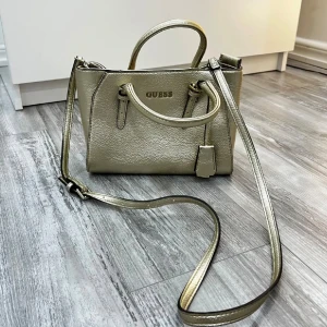 Guess guld handväska med axelrem - Snygg handväska från Guess i metallic guld med struktur och stilrena detaljer. Väskan har både handtag och justerbar axelrem, samt dekorativt hängande tag. Perfekt storlek för det viktigaste och med Guess-logga framtill.