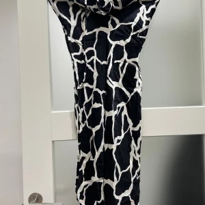 Svartvit mönstrad långklänning - Säljer en svartvit långklänning med coolt grafiskt mönster som påminner om giraff. Klänningen har en lös passform och är ärmlös med en snygg draperad krage. Materialet känns glansigt och lätt, perfekt för dig som gillar att sticka ut med din stil.